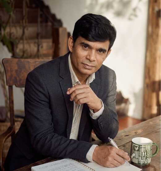 Magician Sooraj, professional mentalist, seated at a wooden table, thoughtfully holding a pen with a notebook and coffee mug in a warm, refined setting.